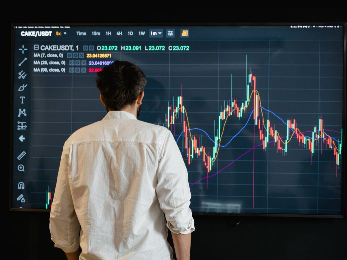 A man in a white shirt stands with his back to a large screen showing a candlestick price chart.