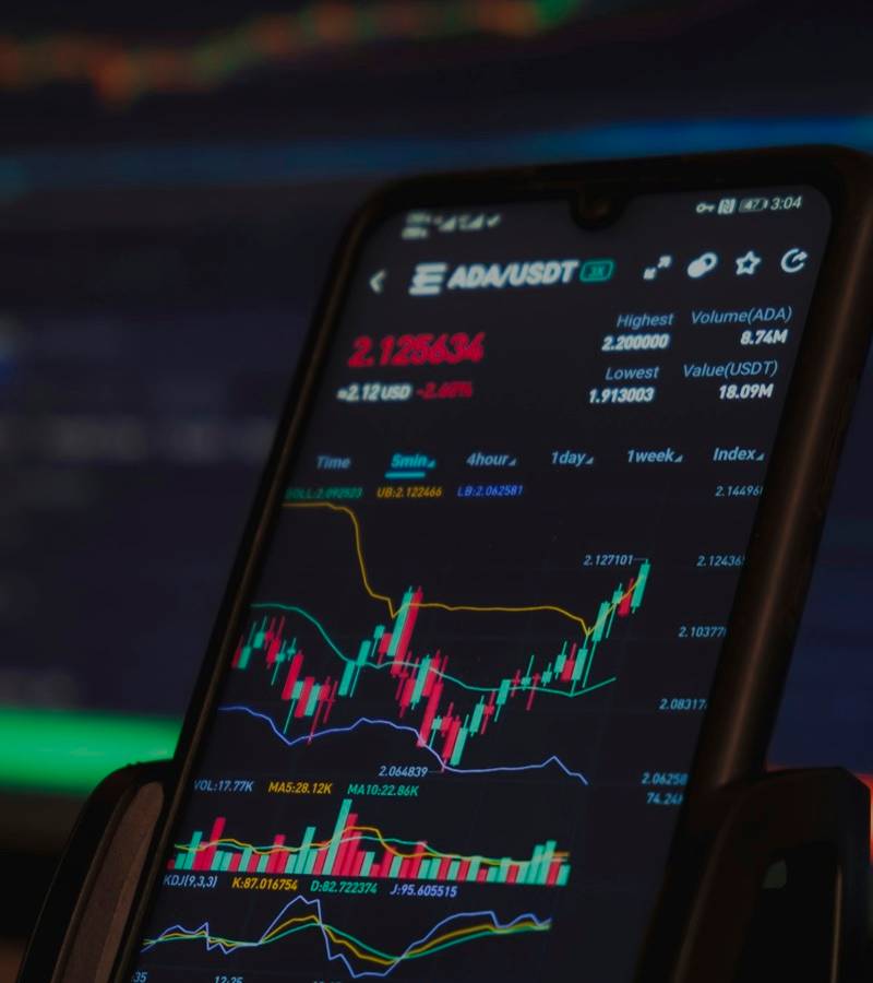 A smartphone on a stand shows a crypto price chart with candlesticks in front of a blurred trading screen.