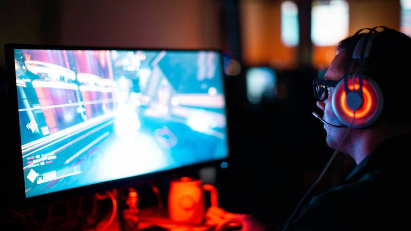 A gamer in an orange-lit headset plays a fast shooter on a monitor in a dark room.
