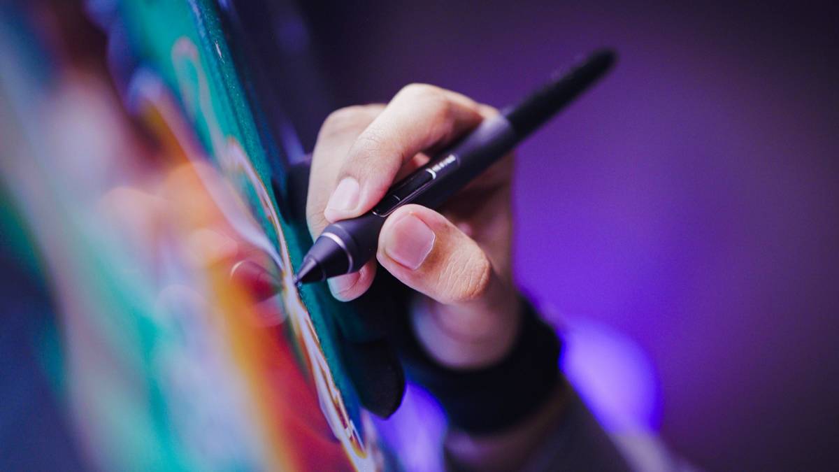 A hand holds a stylus against a colorful digital artwork on a drawing screen, lit in purple.
