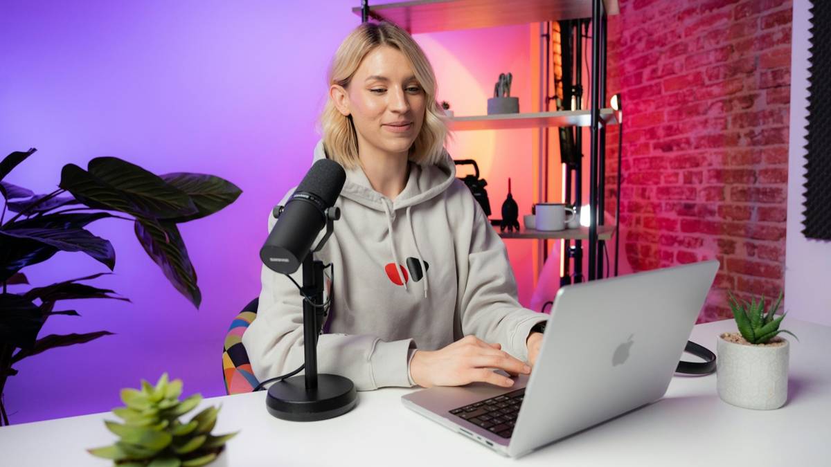 A woman streaming at a desk with a microphone and a laptop in a colourful studio.