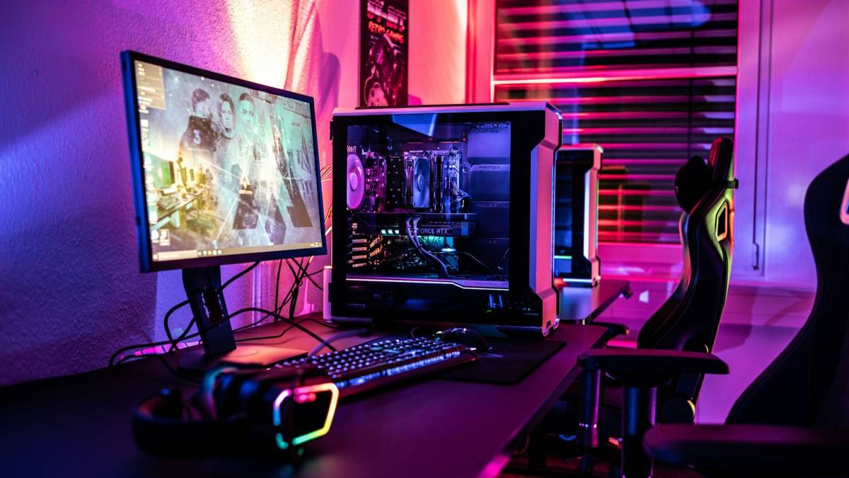 A gaming desk with a monitor, a tower PC and a gaming chair lit in pink and purple.