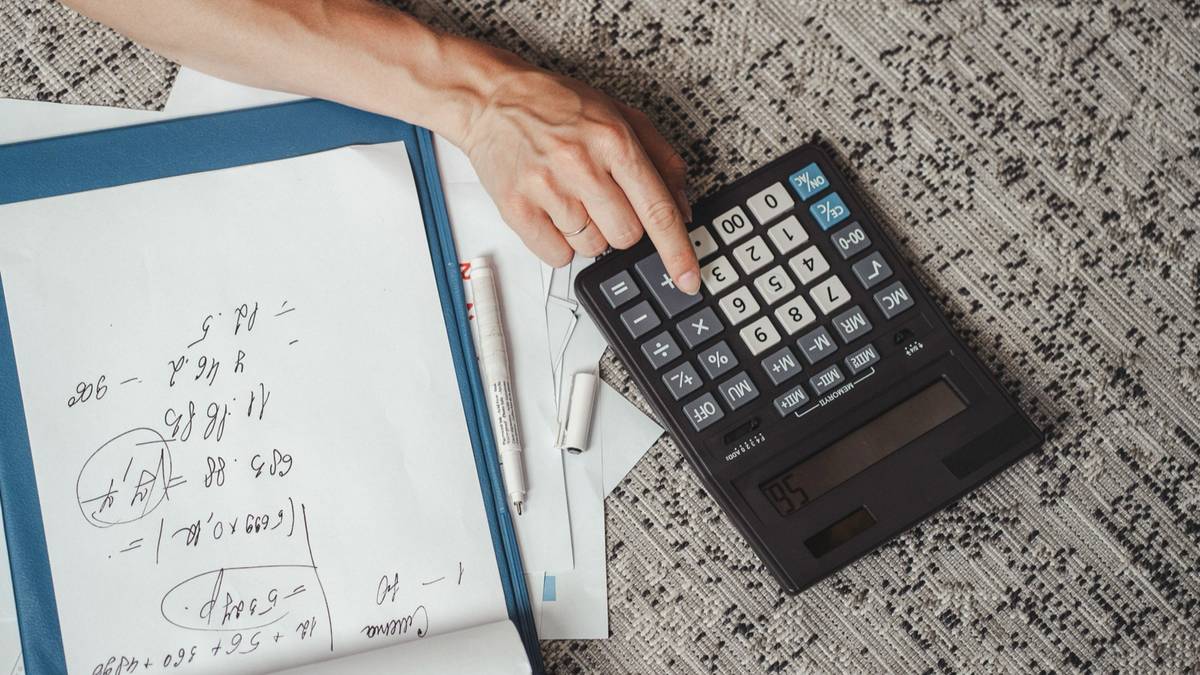 A hand pressing a desk calculator beside a page of handwritten calculations.