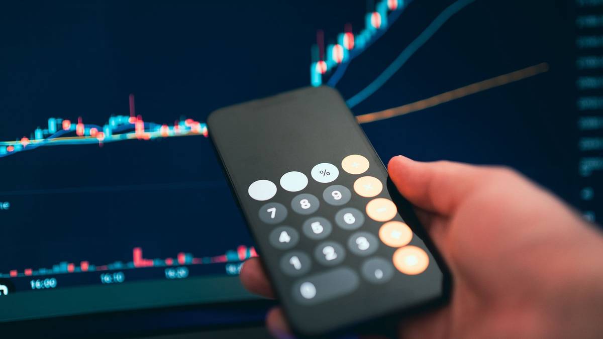 A hand holding a phone with its calculator open in front of a screen of price charts