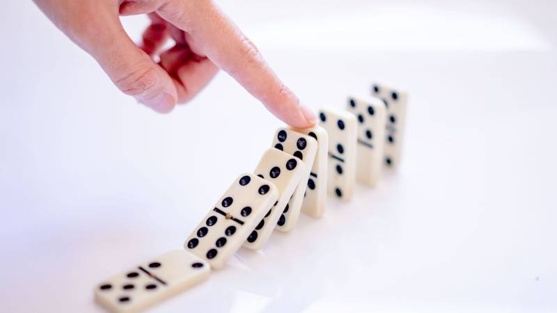 A finger pushes the first of a row of white dominoes that tip over one after another.