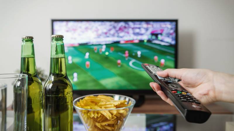 A hand points a remote at a television showing a football match, with beer bottles and a bowl of chips in front.