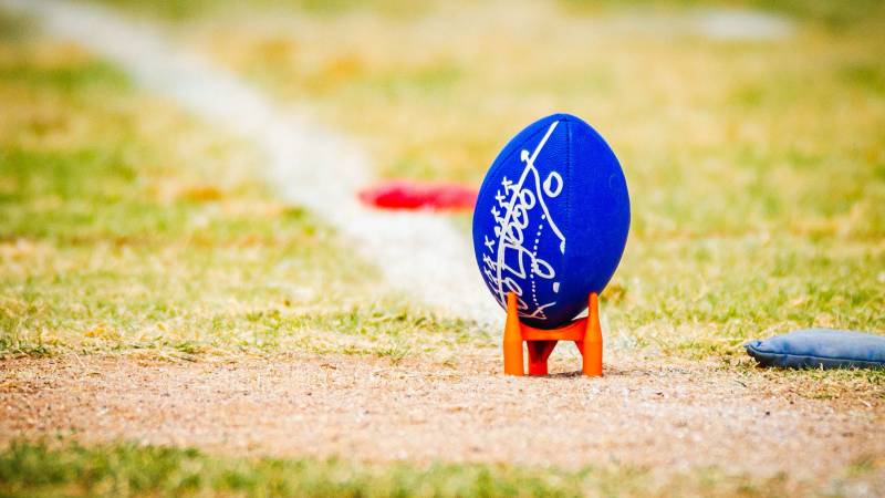 A blue football marked with a hand-drawn play diagram stands on a kicking tee beside a chalk line.