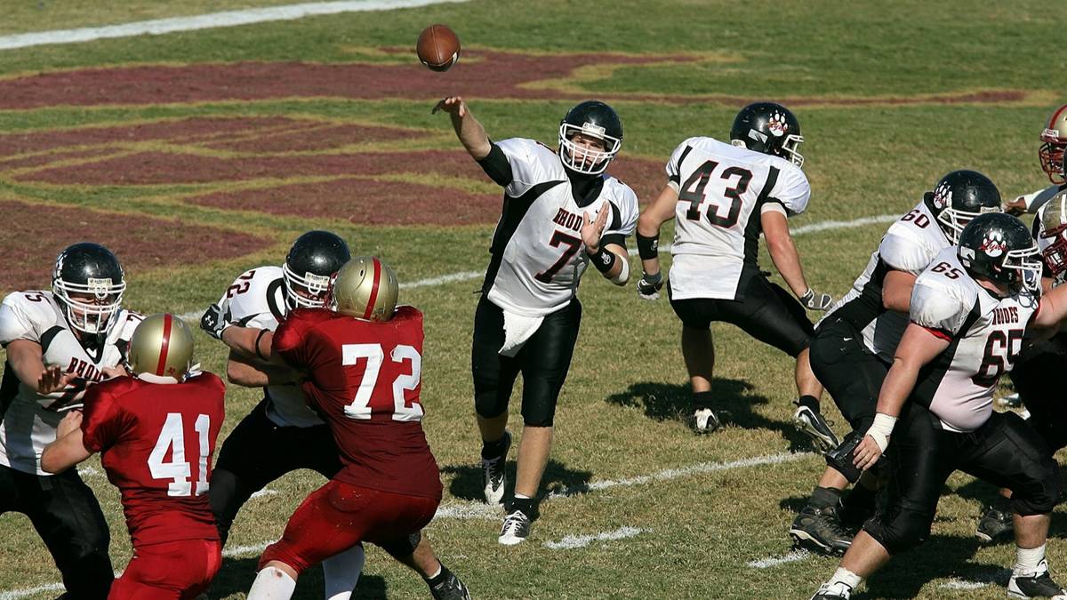 A quarterback throws a pass behind a line of blockers on a grass football field.