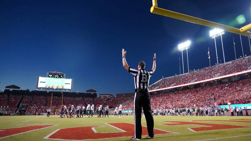 A referee raises both arms under a yellow goalpost as a packed football stadium glows at dusk.