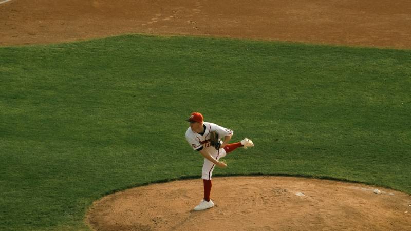 A baseball pitcher in a white and red uniform finishes his delivery from the mound at dusk.