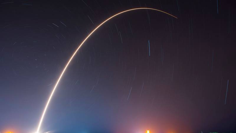 A long exposure of a rocket launch leaving a bright arc across a starry night sky.
