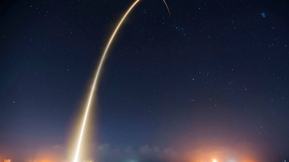 A long exposure light trail of a rocket launch arcs high across a starry night sky.