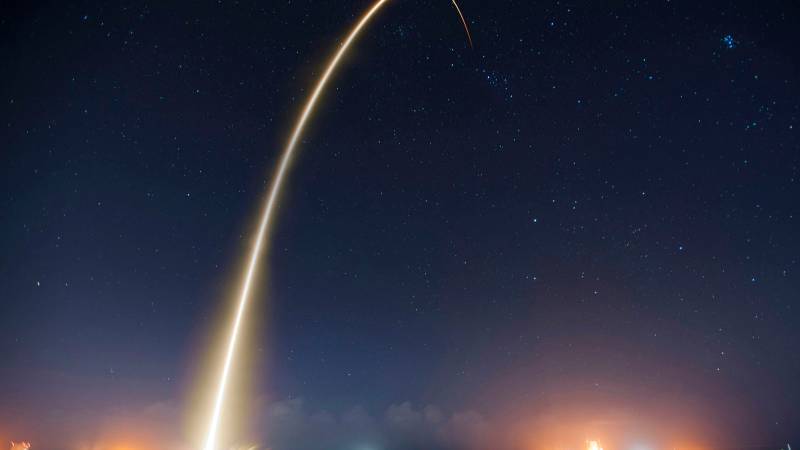 A long exposure light trail of a rocket launch arcs high across a starry night sky.