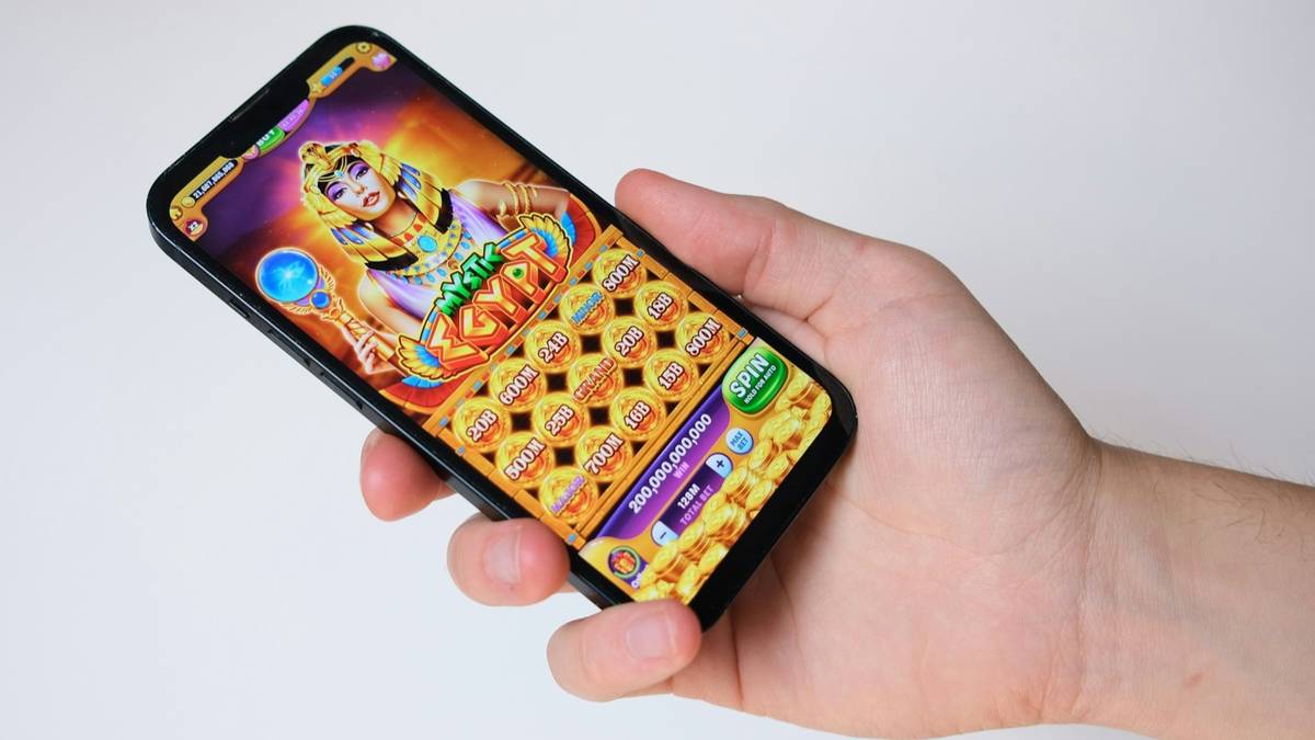 A hand holds a smartphone showing a casino-style game with a grid of gold coin tiles against a plain light background.