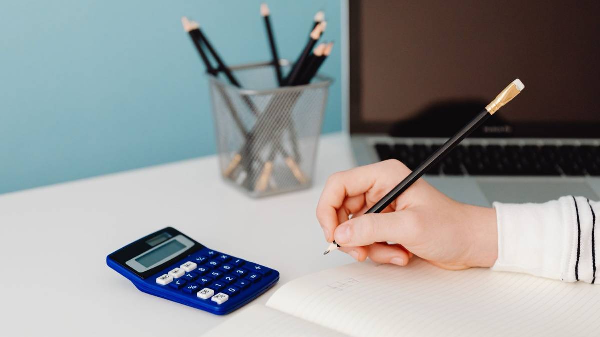 A hand writes with a pencil in a notebook beside a blue calculator and an open laptop on a white desk.