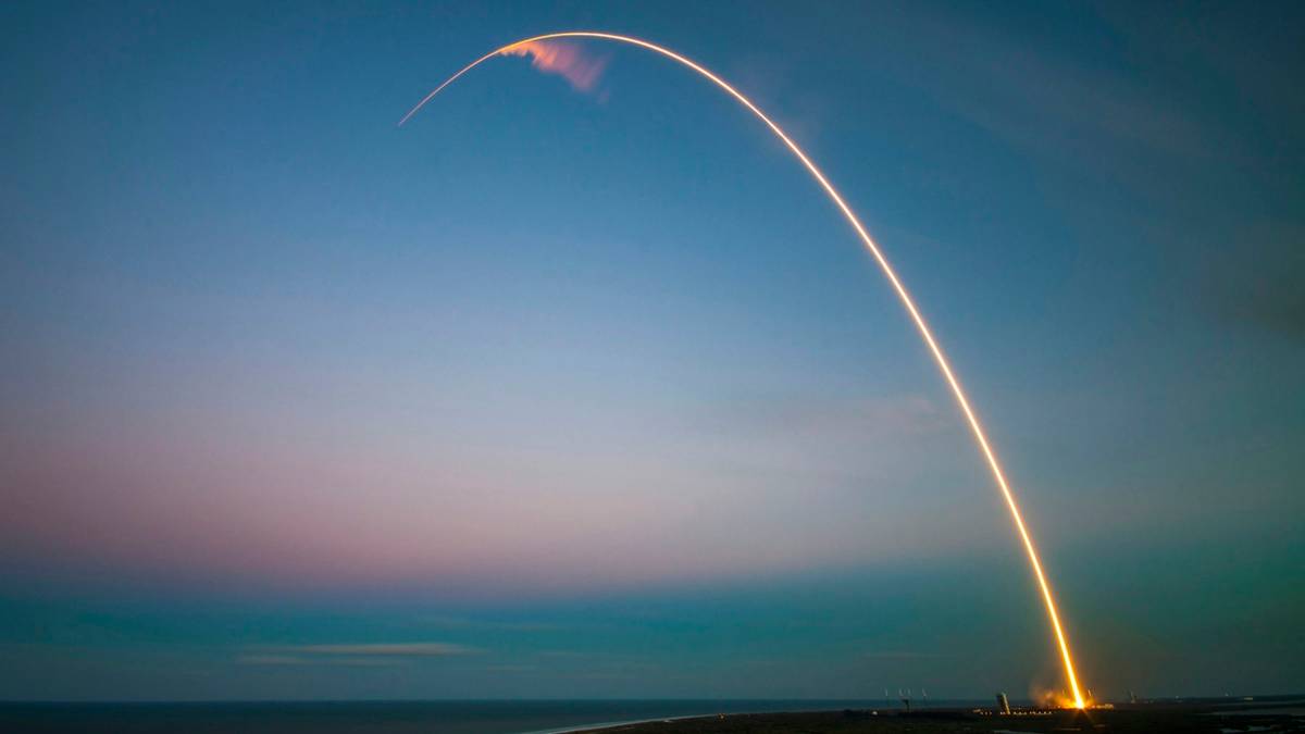 A rocket launch leaves a long glowing arc across a dusk sky over the sea.
