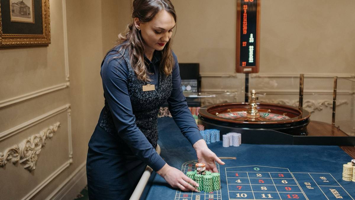 A casino dealer in a navy waistcoat stacks chips on a roulette table with the wheel behind her